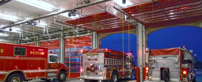 Three fire trucks are parked inside a brightly lit fire station garage with open doors at dusk.