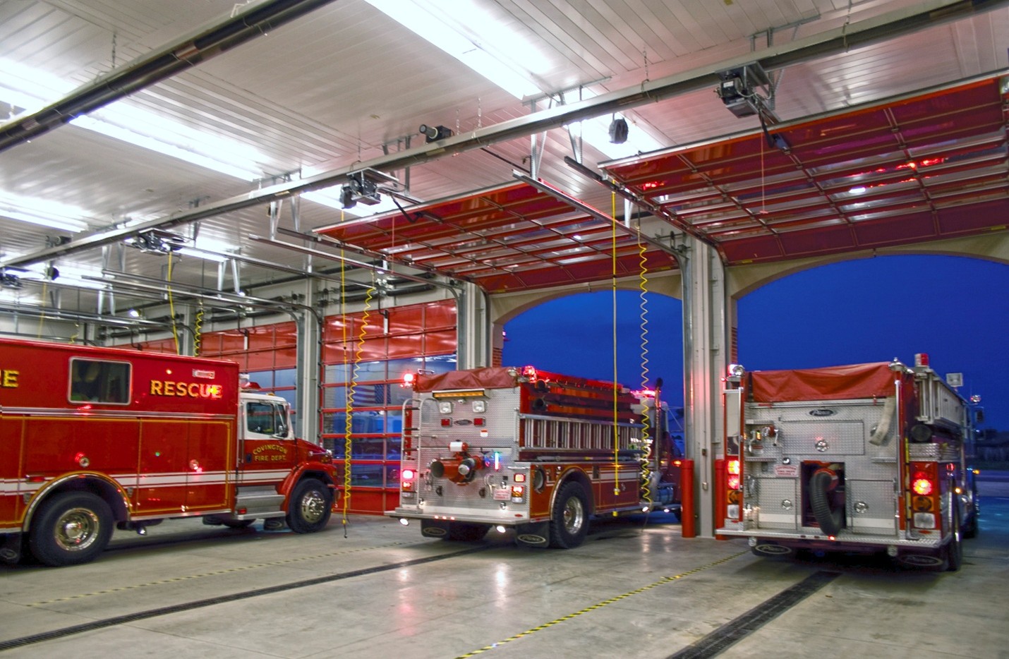 Three fire trucks are parked inside a brightly lit fire station garage with open doors at dusk.