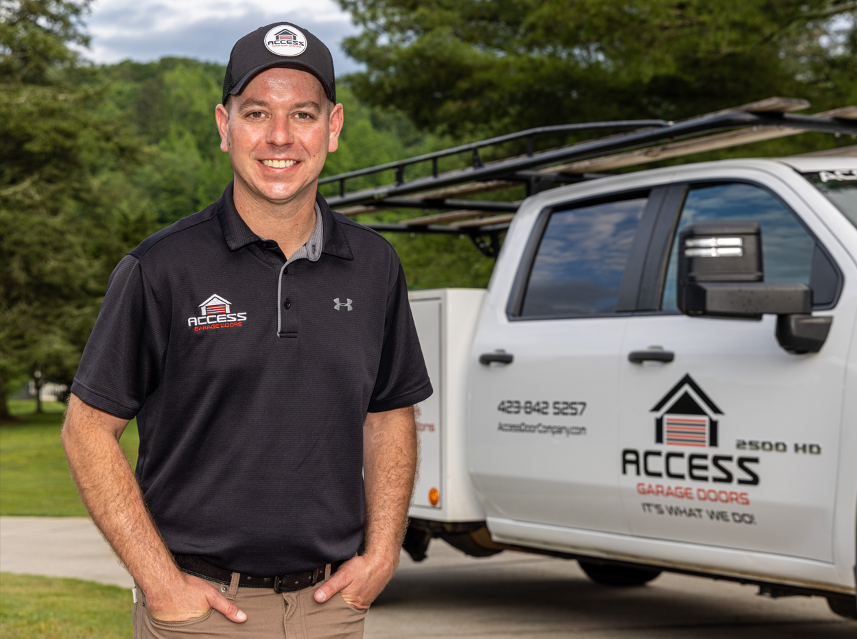A man wearing an Access Garage Doors uniform and cap stands next to a branded company truck parked outdoors on a driveway.